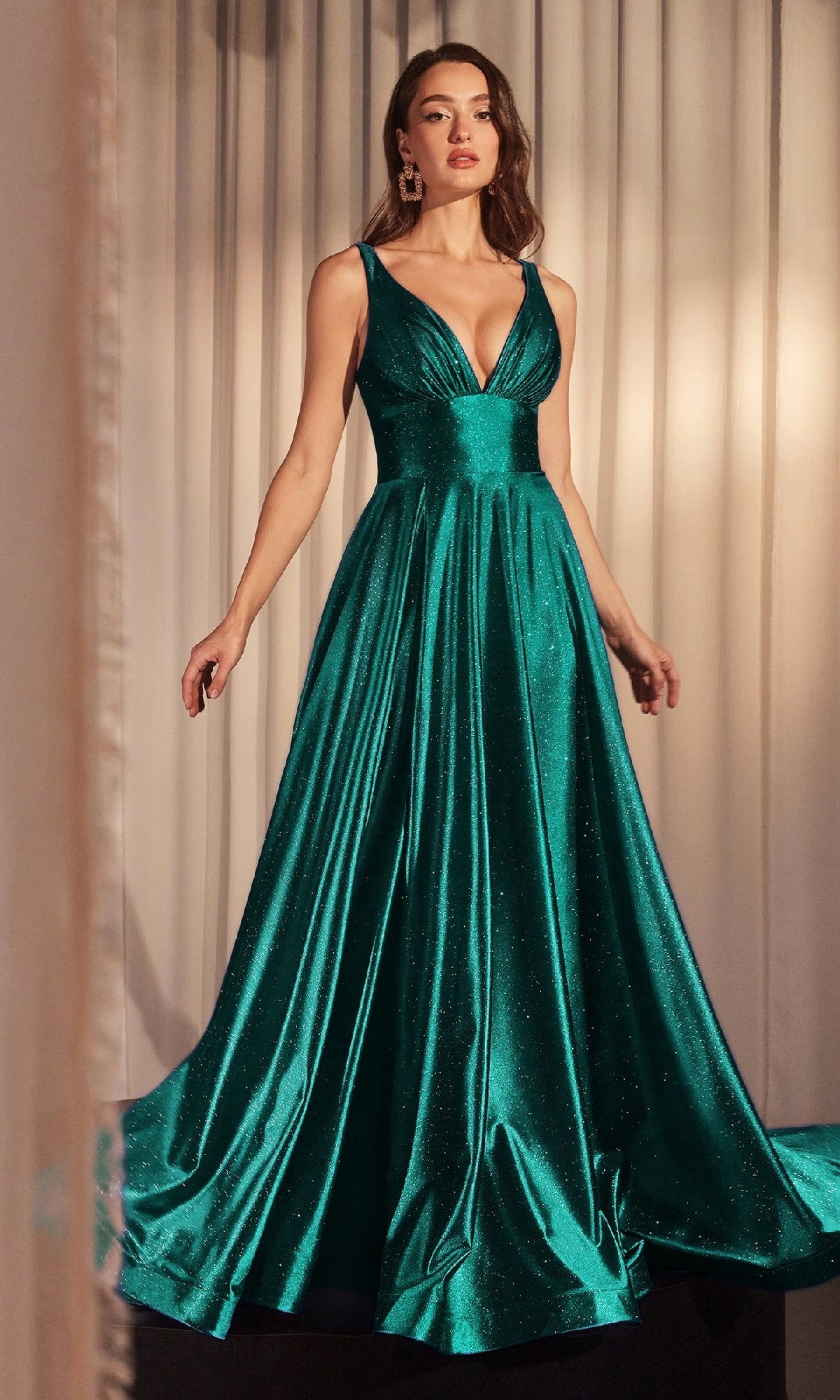 A woman stands before white curtains in the Ladivine CC2349: a sleeveless, deep v-neck, floor-length emerald green glitter prom dress with a full, flowing skirt.